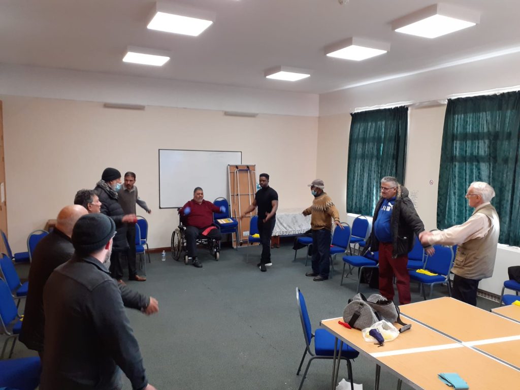 Men standing around the edges of a meeting room, with their arms spread out. One man is sitting in a wheelchair.