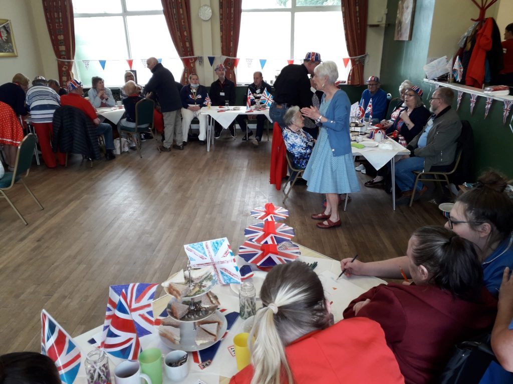 people seated and standing in a church hall decorated with Union Jack bunting