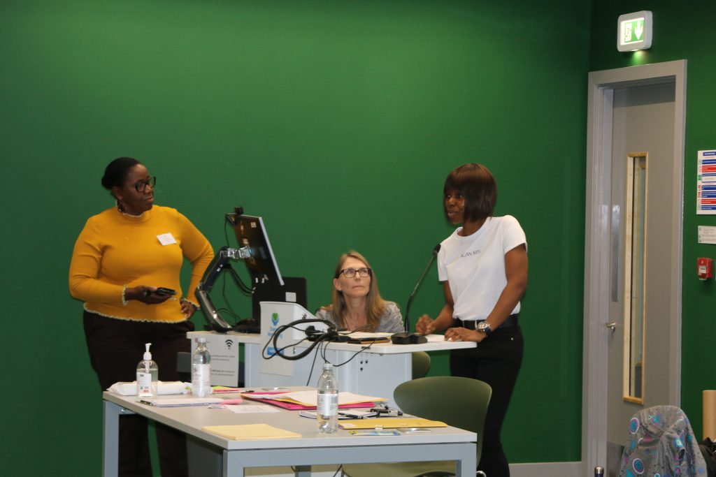 woman standing speaking at a lectern, with 2 women next to her, watching her.