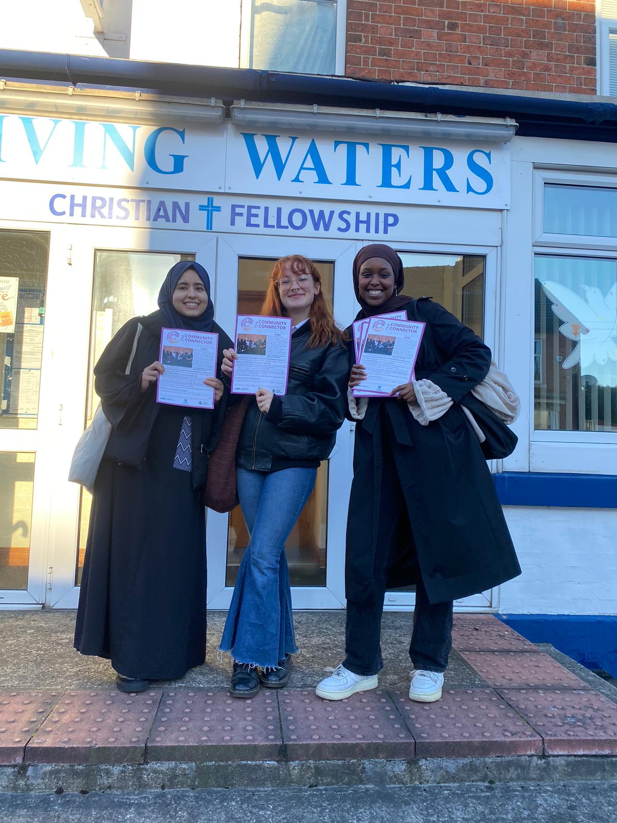 3 women standing outside a building, smiling to camera and holding up community newsletters