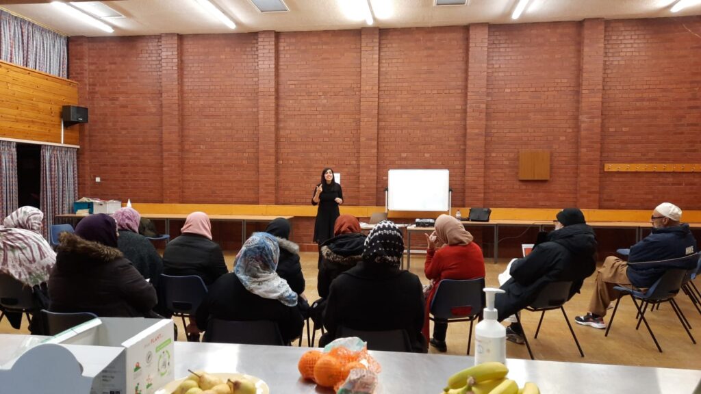 People seated in a community hall, facing away from the camera towards a standing woman who is speaking to them. Fruit on a table in the foreground.