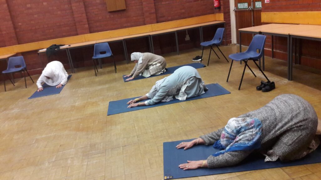 4 women on yoga mats in a community hall, in child's pose.
