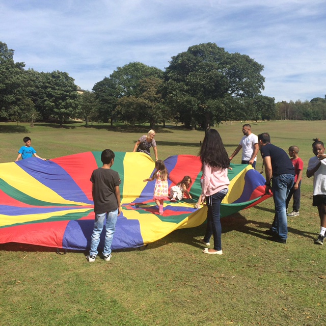 adults and children in a park in the sunshine, playing with a large, colourful parachute