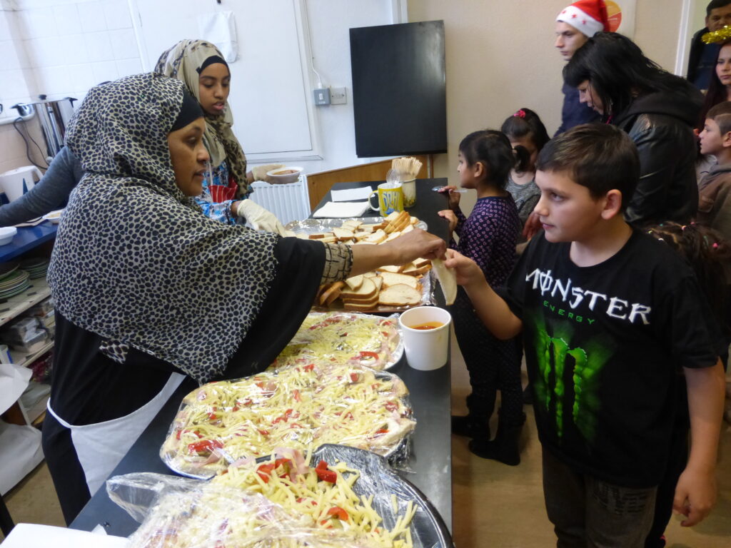 women in headscarves serving behind a table covered in food, with a queue of people at the other side