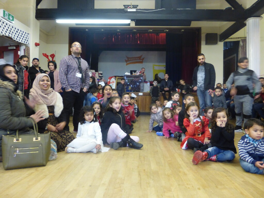 children sitting on the floor of a community hall, looking excited, with adults standing at the edges