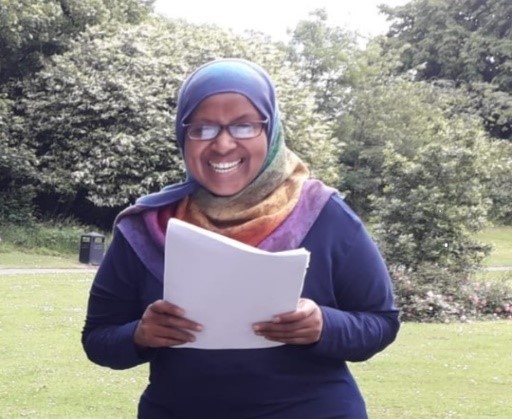 woman in glasses and headscarf standing in a park, smiling to camera