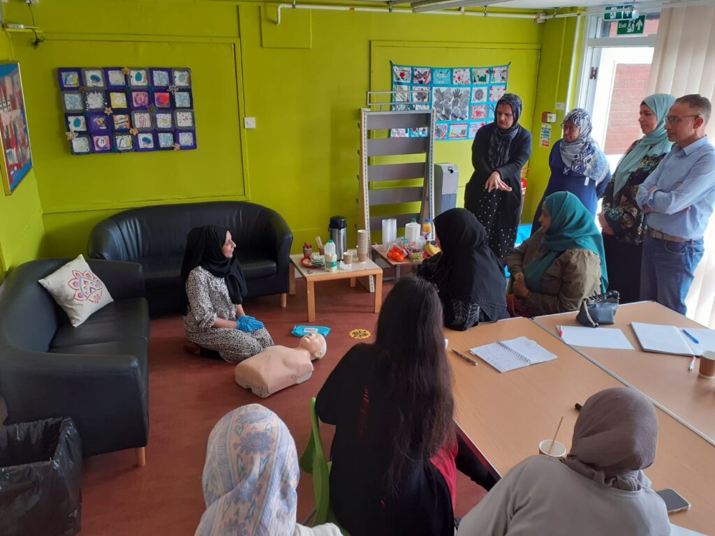 people standing and sitting in a community room, watching a woman with a resuscitation dummy on the floor