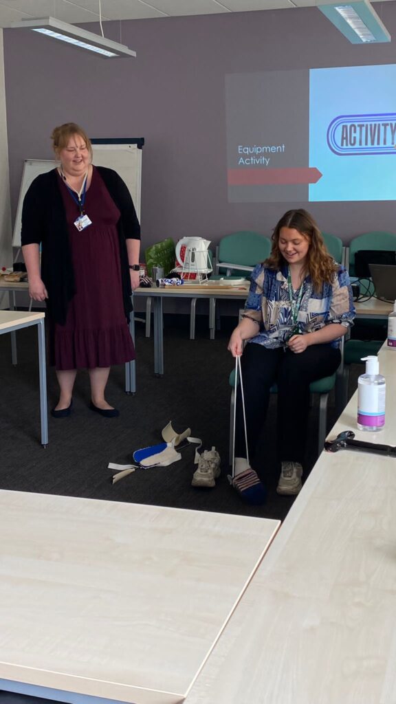 2 female students in a meeting room. One standing watching the other, who is seated and demonstrating how to use an OT aid.