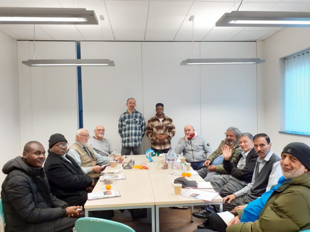 Group of men seated around a large table in a meeting room, with 2 men standing at the end of the table. All are looking towards the camera.