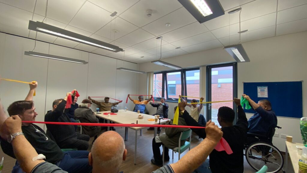 Group of men seated in a meeting room, all holding colourful exercise bands stretched out above their heads.