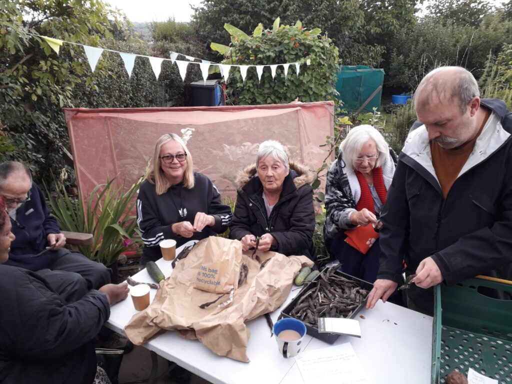 6 people around a table at a community allotment, with produce in paper bags and a tray on the table in front of them