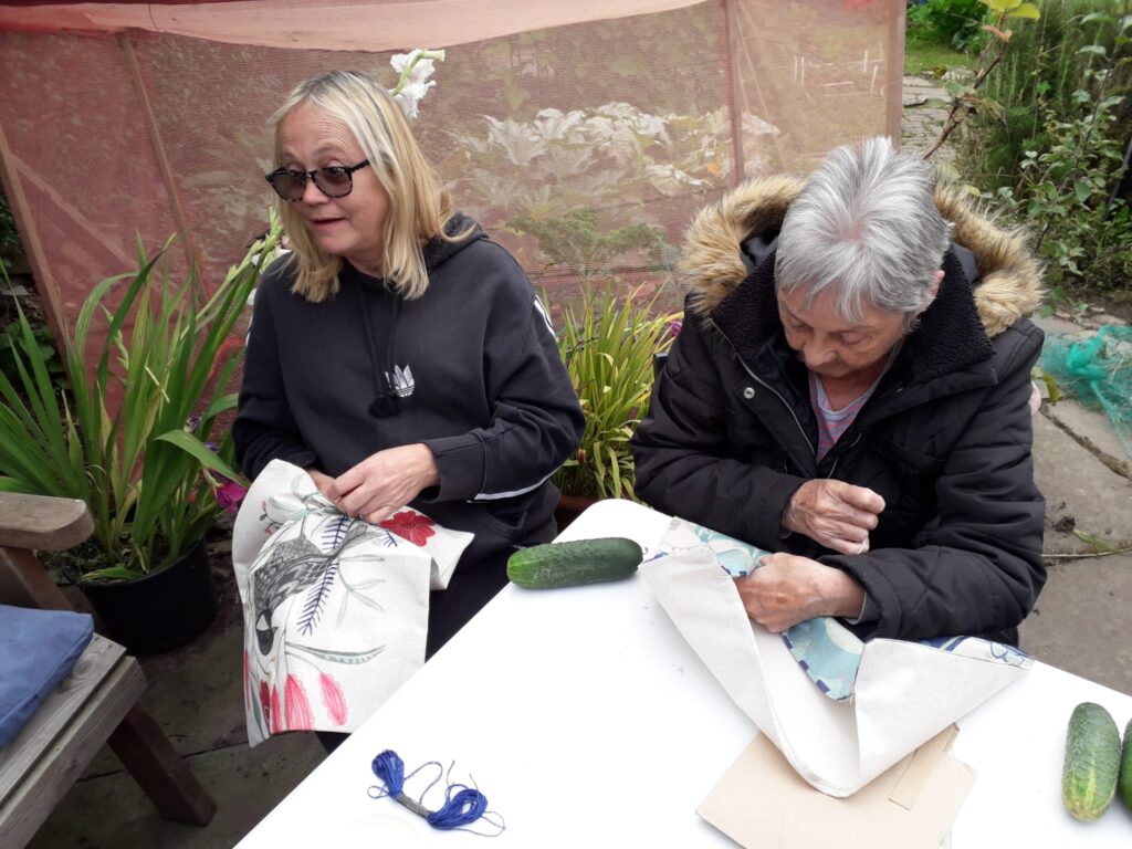 2 women seated outdoors at a table, sewing.