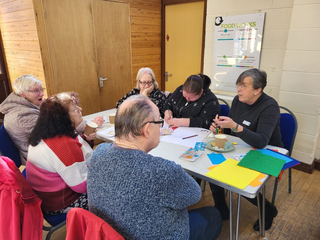 6 people sitting around a table in a church hall with craft materials on the table