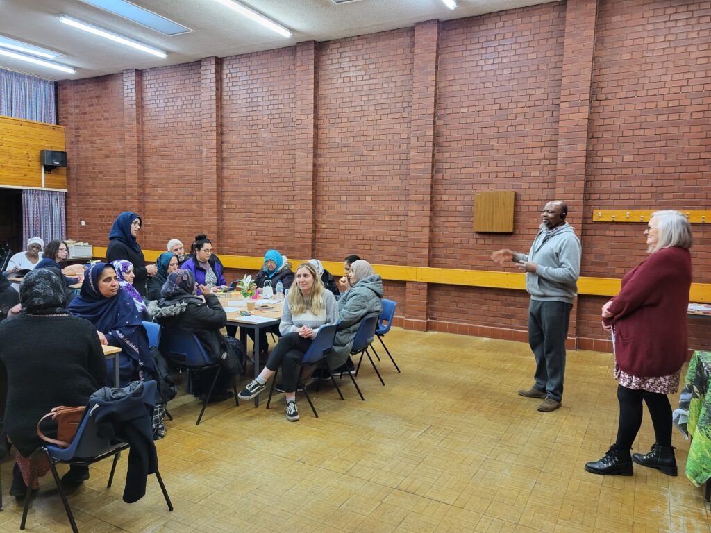 people seated around tables in a community hall, looking at a man and a woman standing at the front