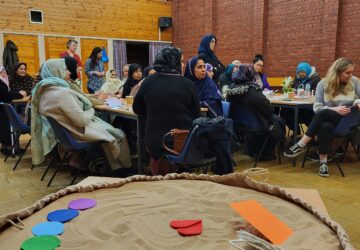 people seated around tables in a community hall, with some craft materials in front of them