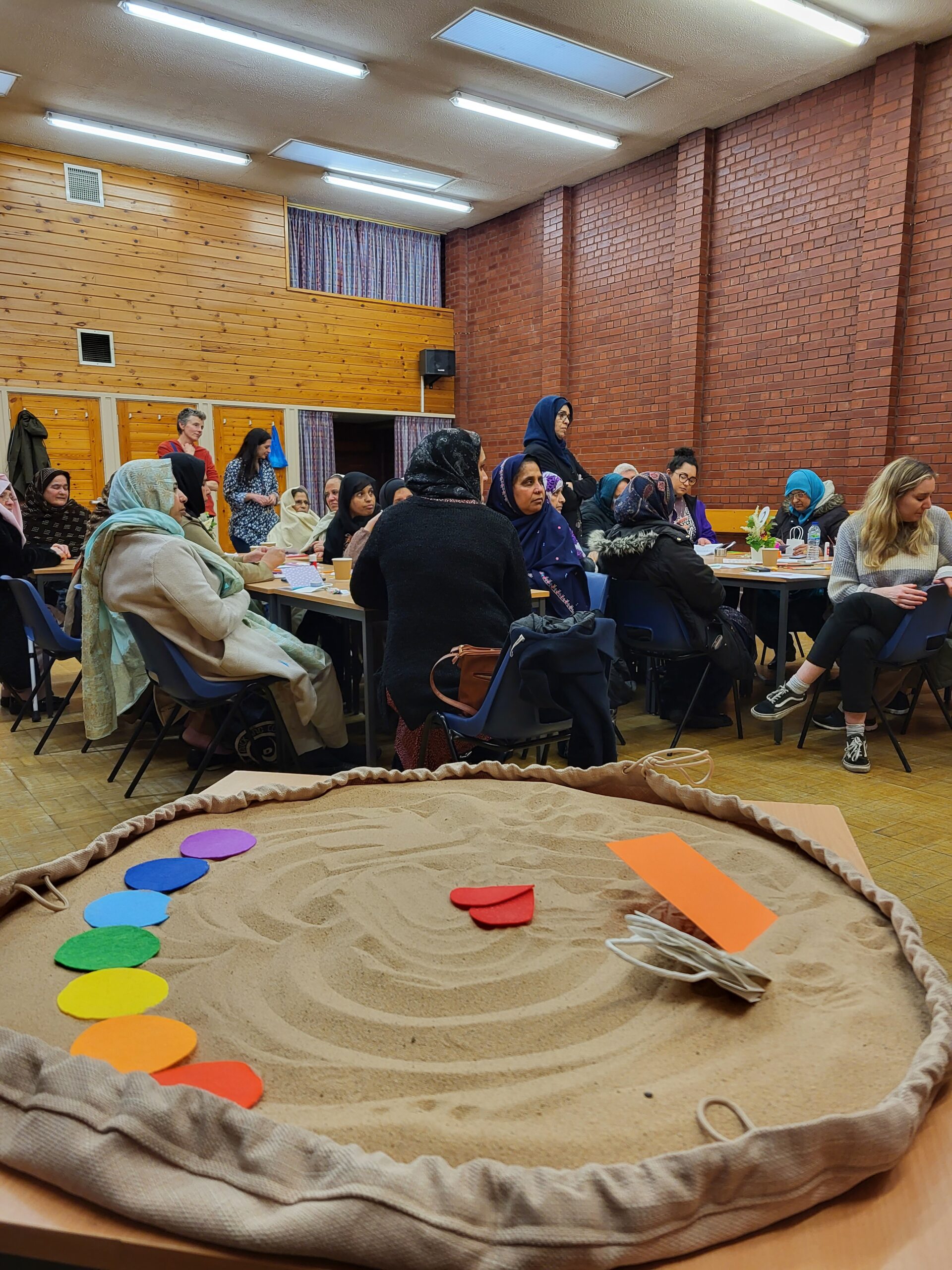 people seated around tables in a community hall, with some craft materials in front of them