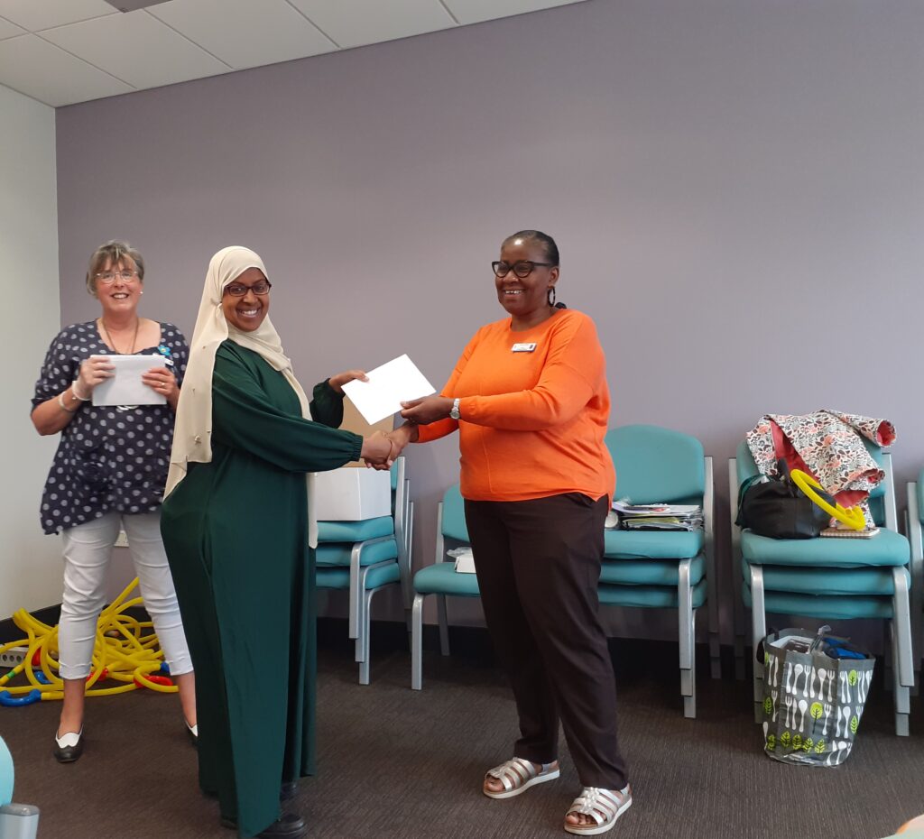 3 women smiling to camera, with 1 handing an envelope to another, whilst shaking hands.