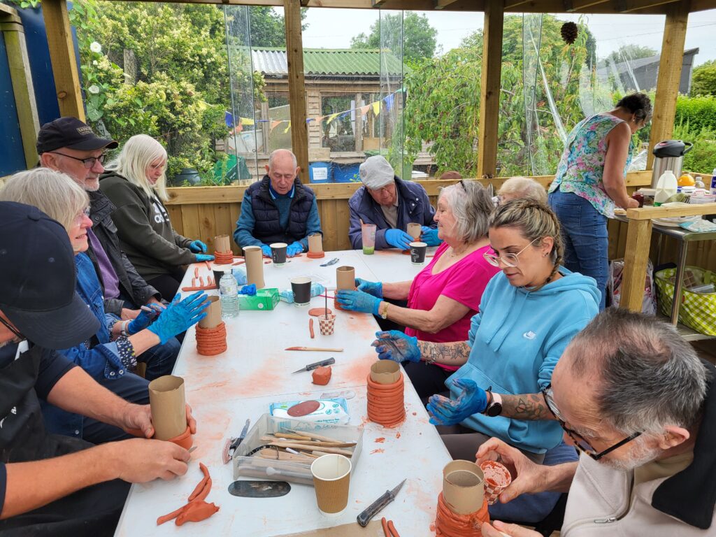 People sitting around a table covered with pottery materials in an outdoor shelter, creating pots together