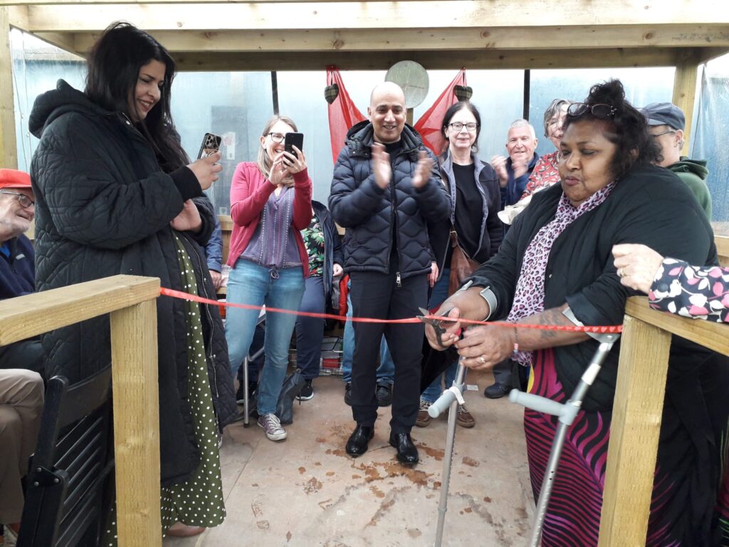 Woman cutting red ribbon at the entrance to an allotment shelter, watched by a group of smiling people