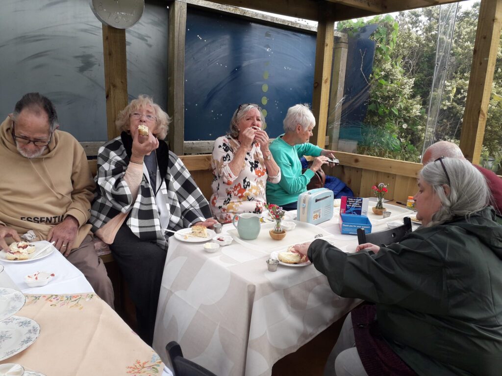 People sitting enjoying a cream tea around a table in an outdoor shelter