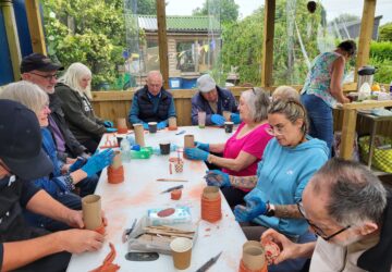 large group of people sitting around a table in a community allotment shelter, making clay models