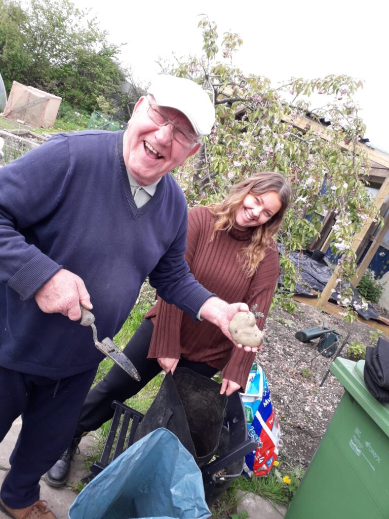 Man and woman laughing and looking to camera whilst standing outside at a community allotment. Man is holding a potato and a trowel.