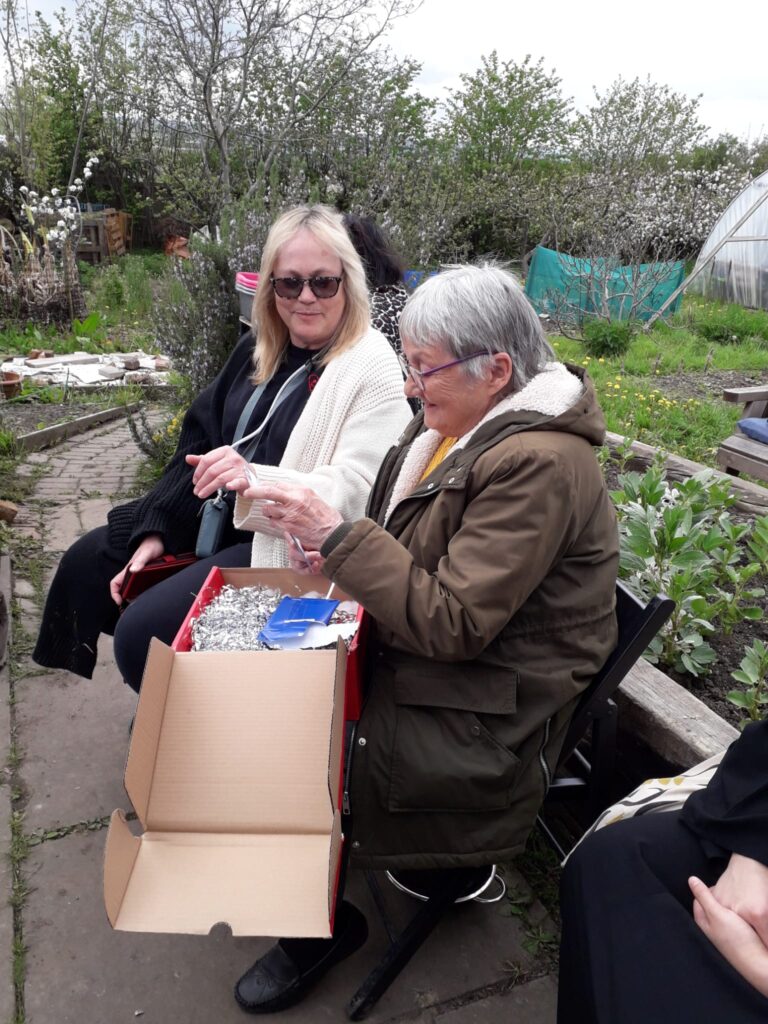 2 women seated outside at a community allotment, looking at items in a box.