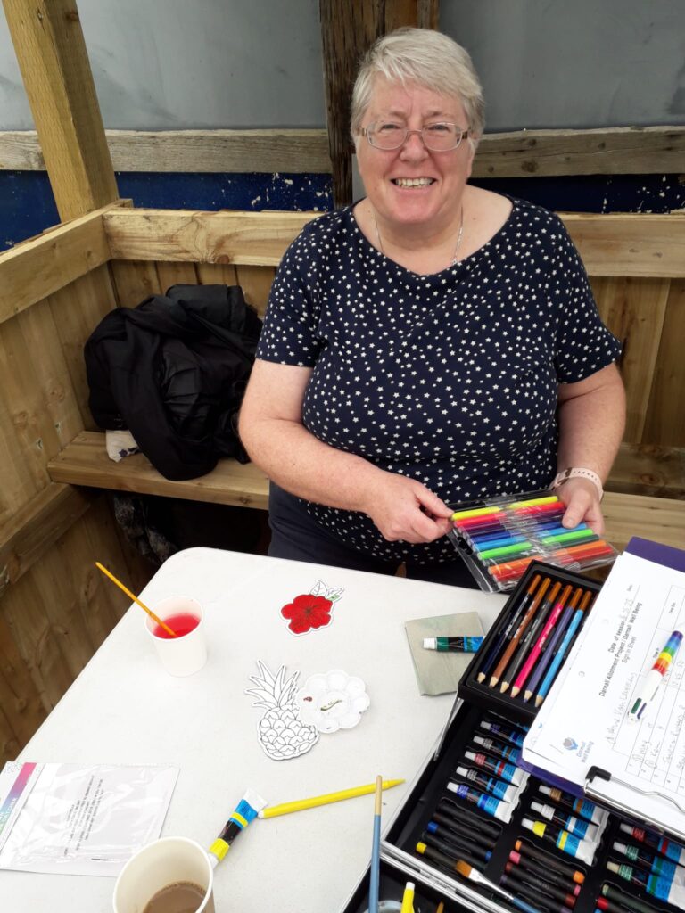 woman seated at a table covered in art materials, smiling to camera