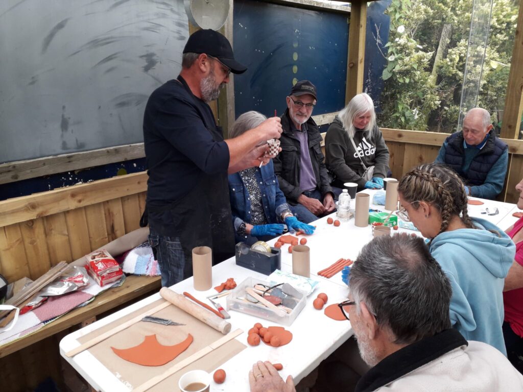 group of people at a table making clay models, whilst a man stands up to demonstrate what to do