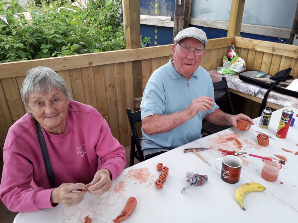 a man and woman sitting in a community allotment shelter, smiling to camera whilst making clay models