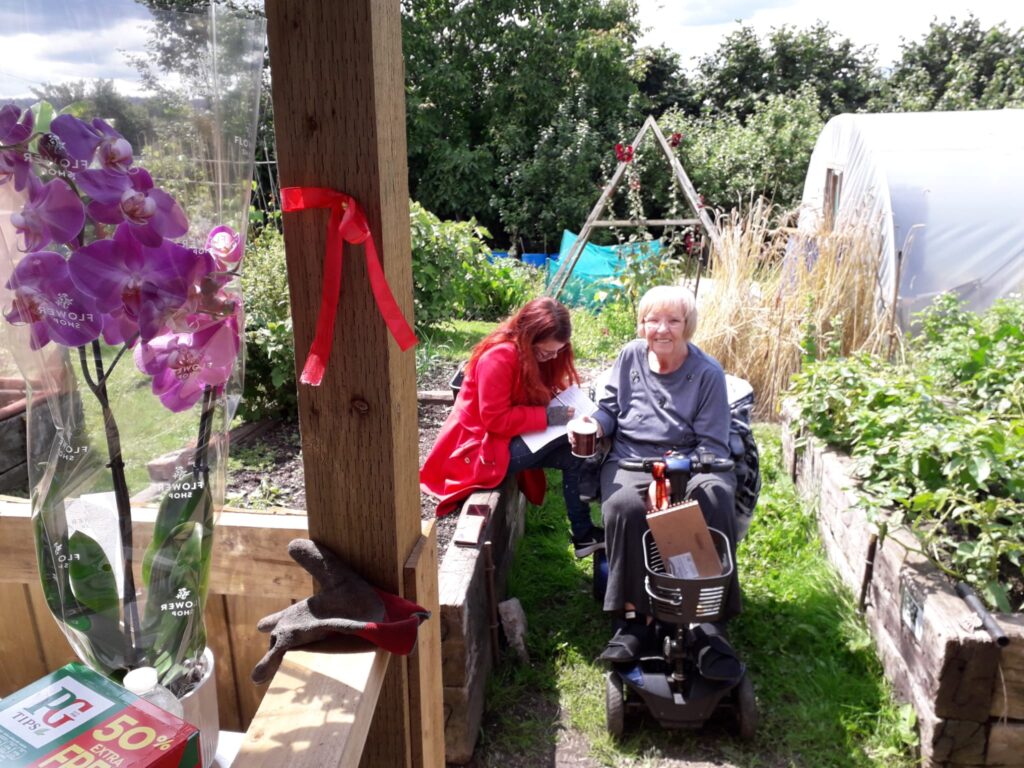 woman in mobility scooter on a community allotment in the sunshine, smiling to camera, with another woman sitting next to her on a raised bed.
