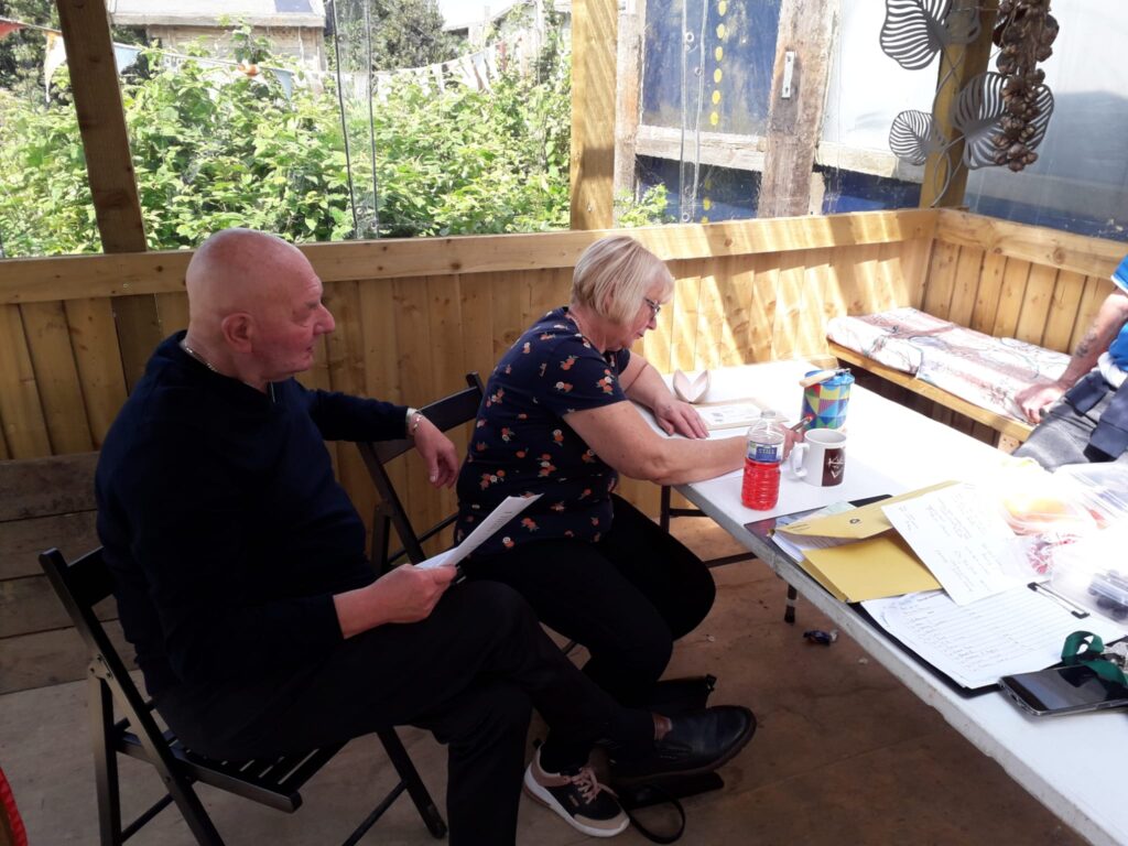2 people sitting in a community allotment shelter, with pieces of paper